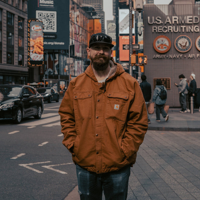 Tyler in a brown jacket standing on a city street with tall buildings and advertisements in the background.